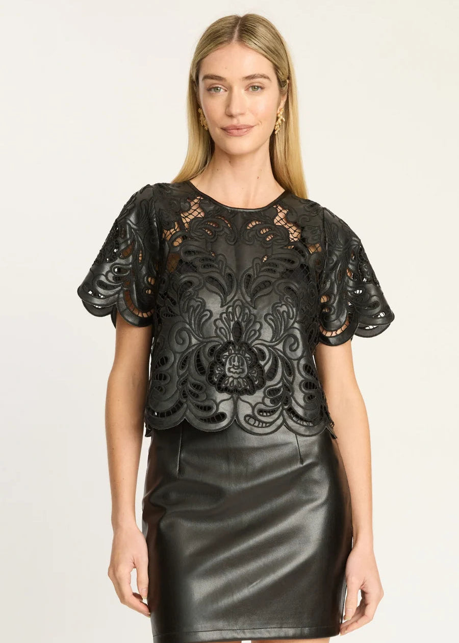 A woman modeling a black short-sleeve top with intricate floral lace applique and a black faux-leather skirt against a plain light background.