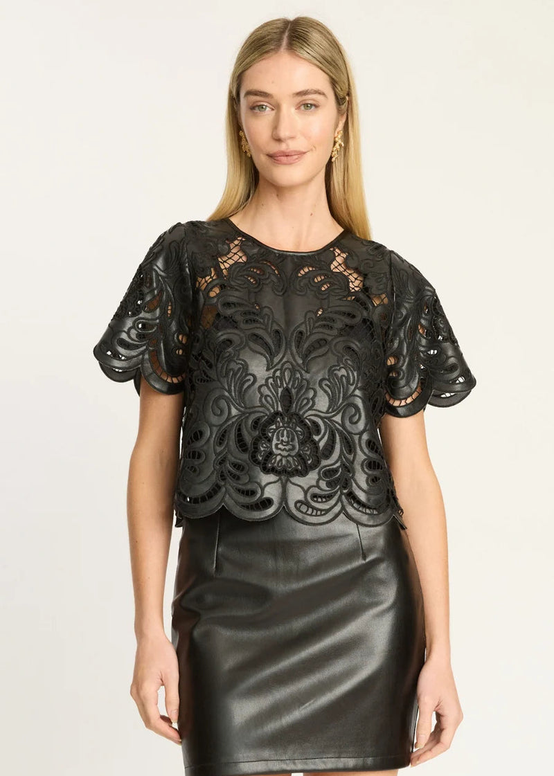 A woman modeling a black short-sleeve top with intricate floral lace applique and a black faux-leather skirt against a plain light background.
