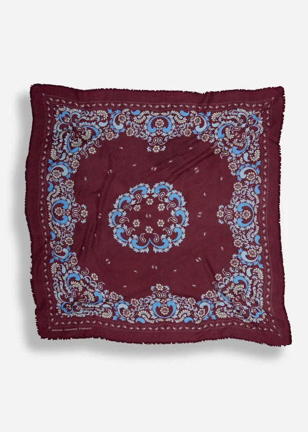 A square bandana scarf in deep burgundy, featuring a central medallion and ornate paisley borders in light blue and cream, laid flat against a light gray background.