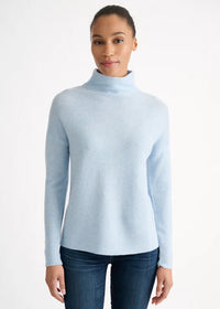 A woman wearing a light blue ribbed mock neck sweater and dark blue jeans, standing against a plain white studio background.