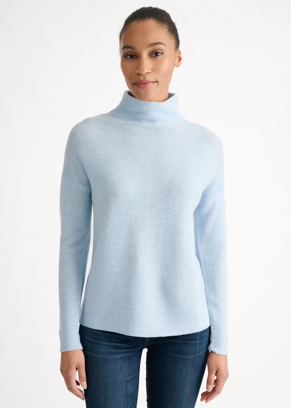 A woman wearing a light blue ribbed mock neck sweater and dark blue jeans, standing against a plain white studio background.