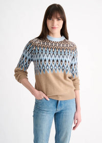 A woman modeling a beige knit sweater with a blue, brown, and white geometric pattern across the chest and shoulders, paired with blue jeans, against a plain white background.
