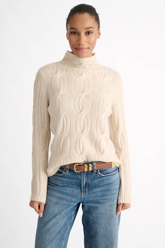 Woman wearing a beige cable knit sweater and blue jeans on a white background