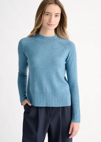 A woman wearing a light blue knit crewneck sweater and dark navy trousers, standing against a plain white studio background.
