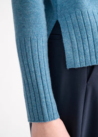 Close-up of a person wearing a blue ribbed knit sweater with a side slit and a dark navy skirt or dress, against a plain light background.