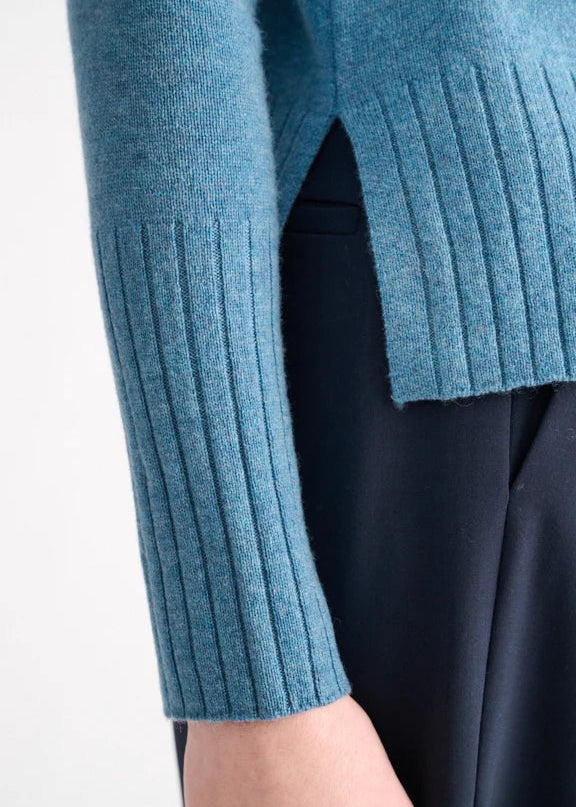 Close-up of a person wearing a blue ribbed knit sweater with a side slit and a dark navy skirt or dress, against a plain light background.