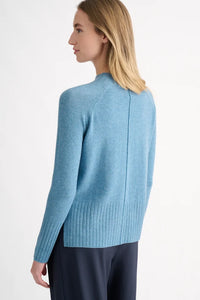 A woman shown from the back wearing a light blue knit sweater with a ribbed hem and dark navy pants, photographed against a clean white studio background.
