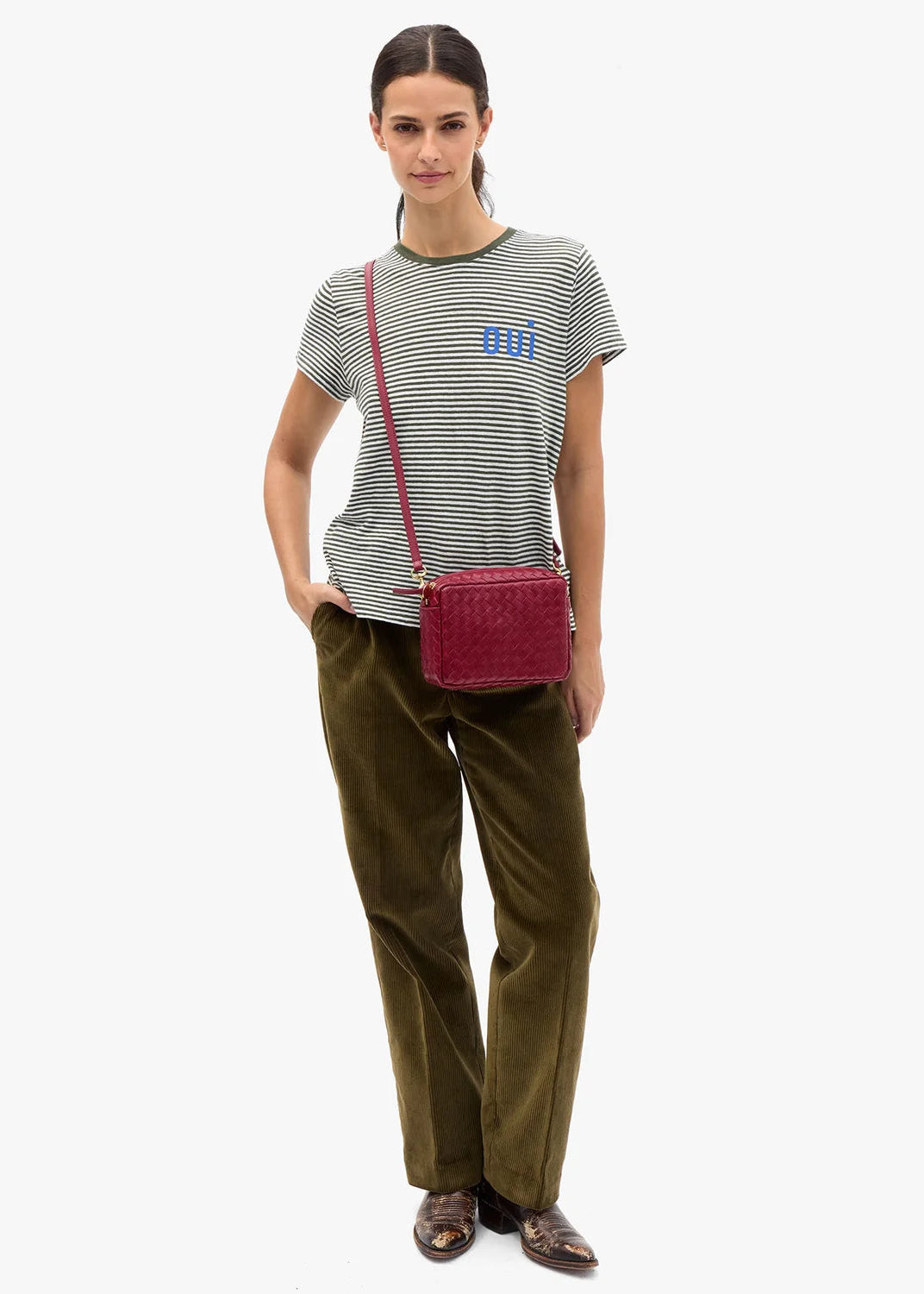 A model stands against a light gray studio background wearing a green and white striped t-shirt with a blue logo, olive green trousers, and brown leather loafers, while carrying a dark red woven crossbody bag.