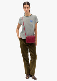 A model stands against a light gray studio background wearing a green and white striped t-shirt with a blue logo, olive green trousers, and brown leather loafers, while carrying a dark red woven crossbody bag.
