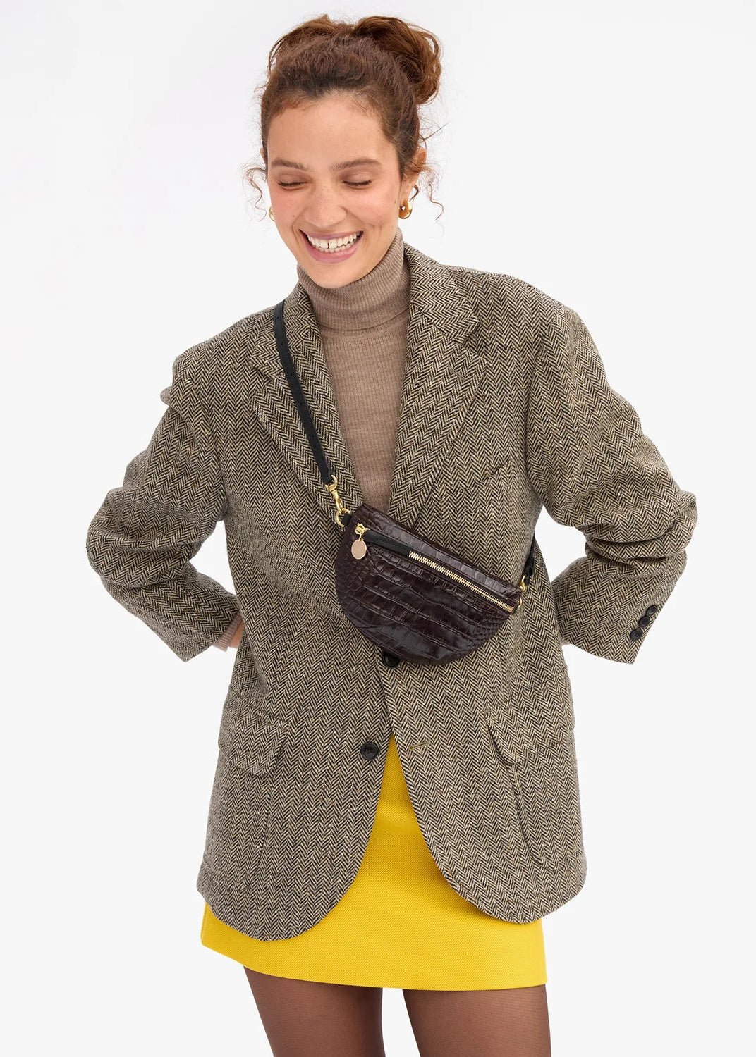 A woman wearing a brown tweed blazer over a brown turtleneck and a yellow skirt, carrying a dark brown crocodile-embossed crossbody bag with gold hardware against a plain white background.
