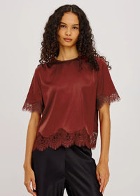 A woman modeling a short-sleeve satin blouse in deep burgundy with lace trim at the hem and sleeve cuffs, paired with black satin pants, against a light gray studio background.