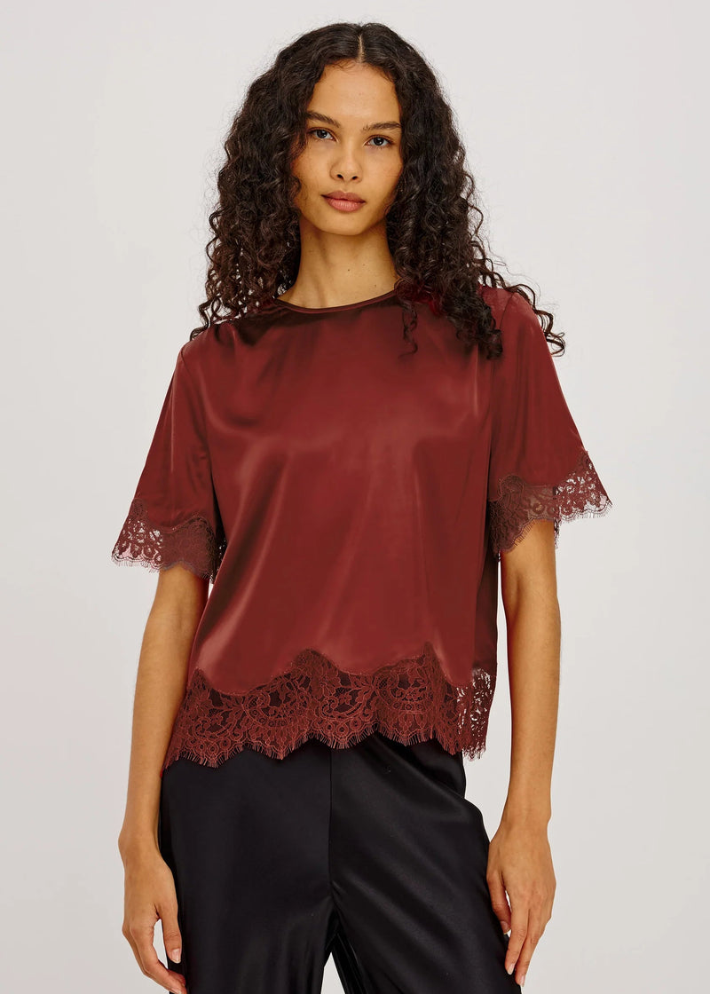 A woman modeling a short-sleeve satin blouse in deep burgundy with lace trim at the hem and sleeve cuffs, paired with black satin pants, against a light gray studio background.