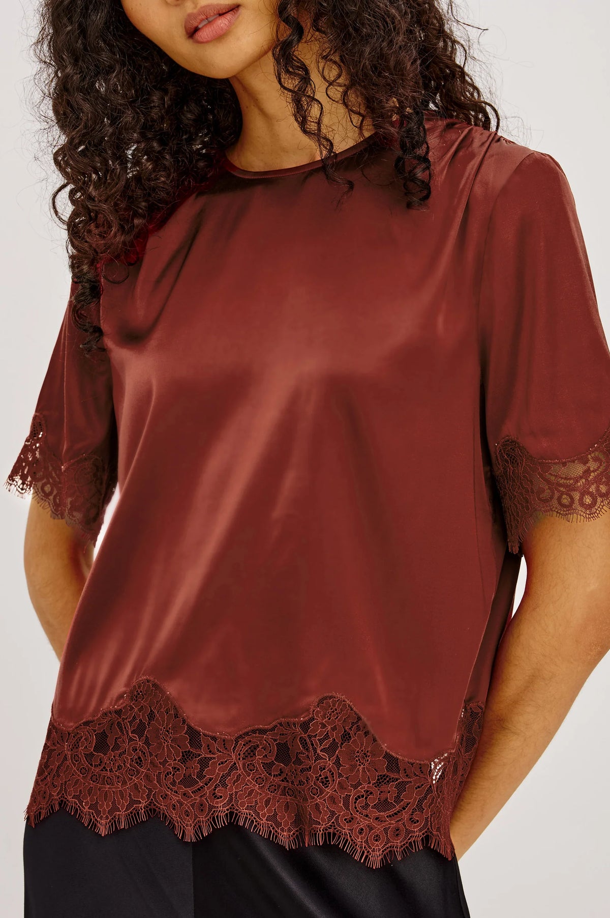 A woman wearing a rust-brown satin short-sleeve top with dark lace trim at the hem and sleeve cuffs, paired with black bottoms, against a light gray studio background.