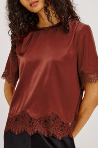 A woman wearing a rust-brown satin short-sleeve top with dark lace trim at the hem and sleeve cuffs, paired with black bottoms, against a light gray studio background.