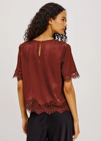 A woman shown from the back wearing a short-sleeve satin blouse in a deep burgundy or rust red color, featuring lace trim at the sleeve cuffs and hem, paired with black pants against a light gray studio background.