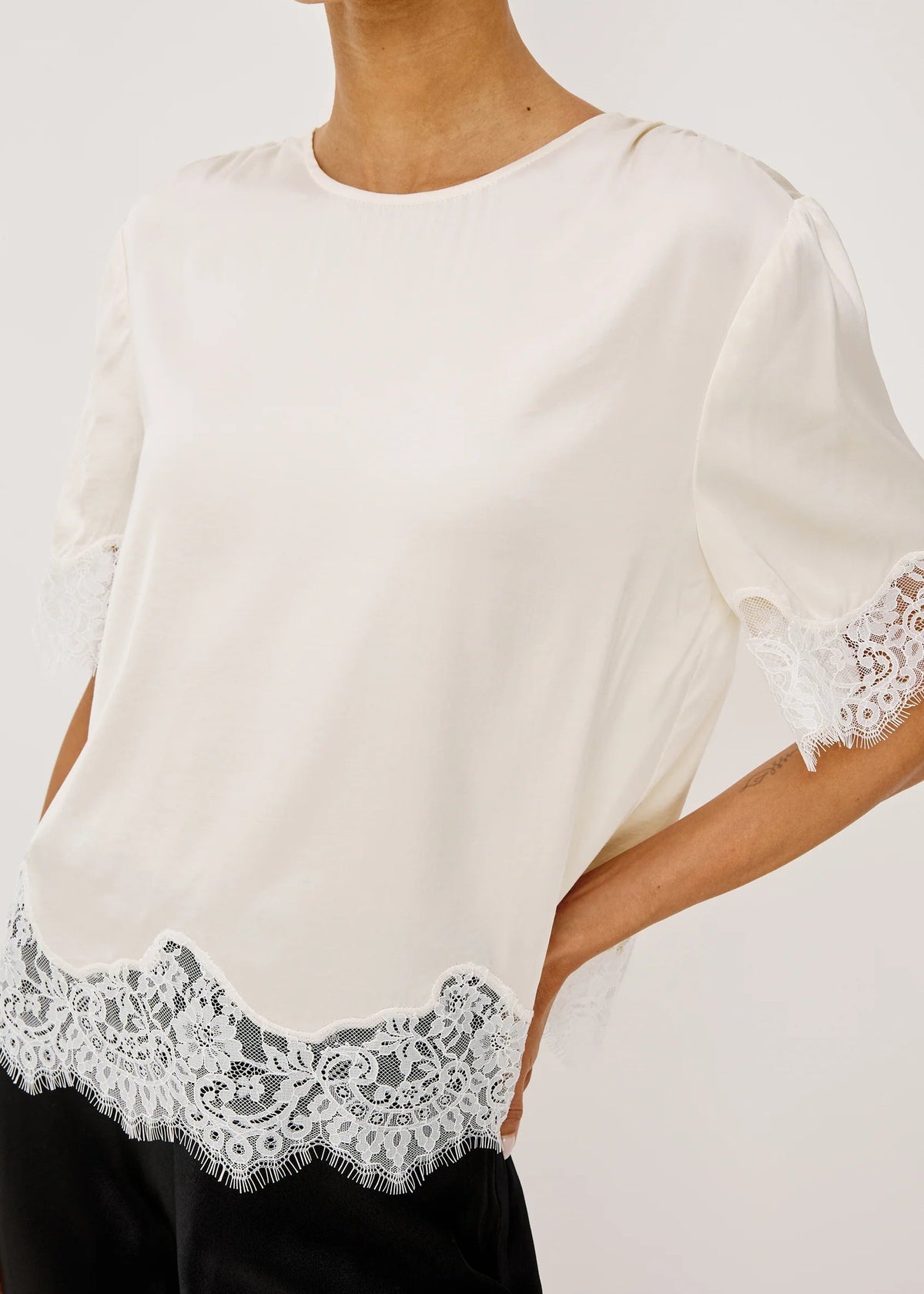 An off-white short-sleeve blouse with delicate white lace trim at the hem and sleeve cuffs, worn by a model against a light gray studio background.