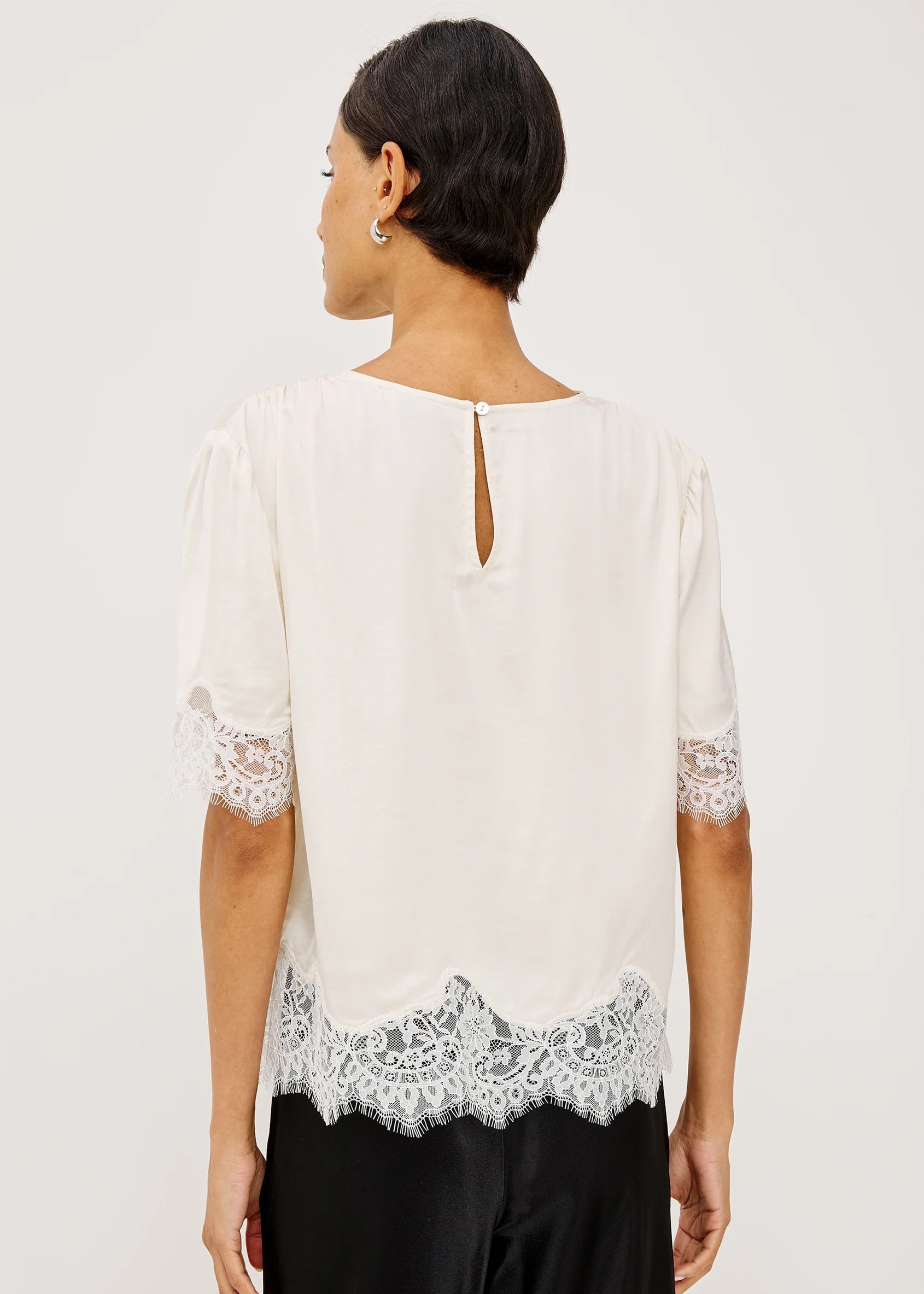 A woman shown from the back wearing a white blouse with short puff sleeves, lace trim at the cuffs and hem, and a small keyhole cutout at the neckline, paired with black pants against a light gray studio background.
