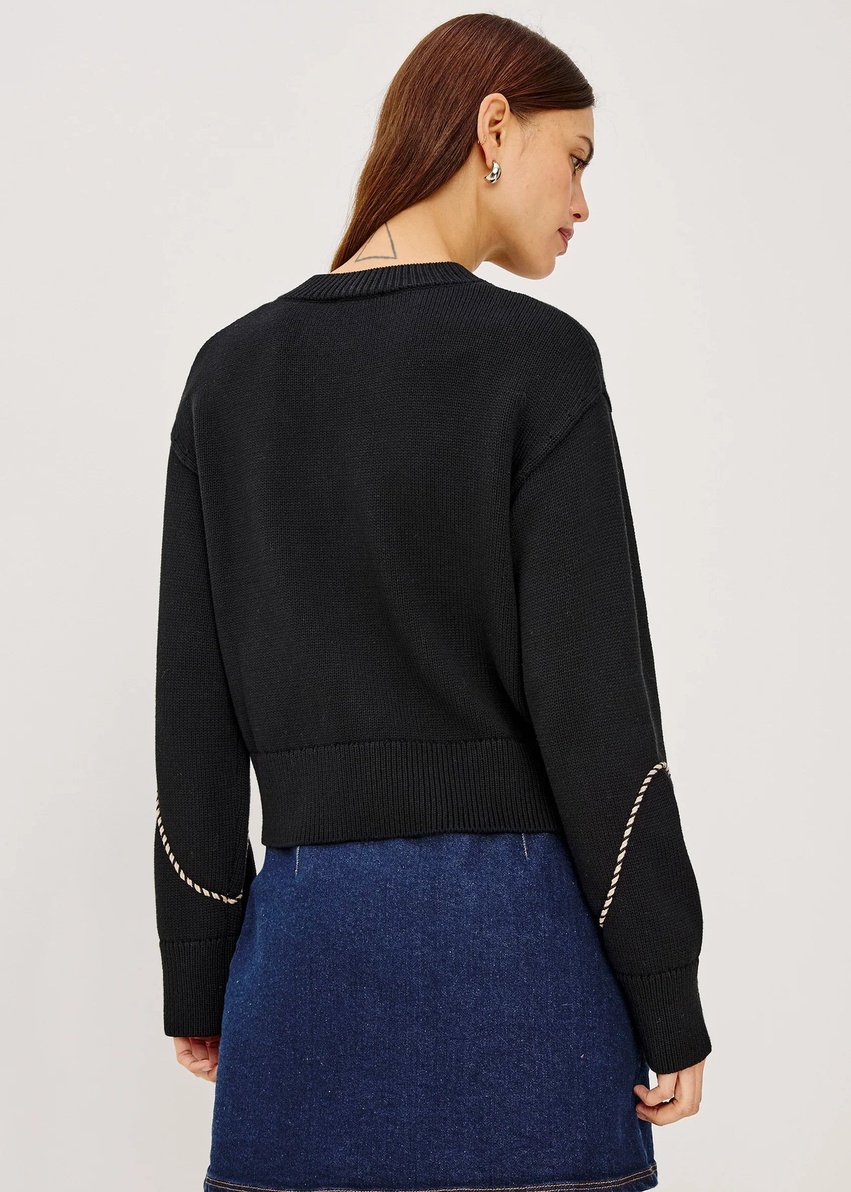 A woman shown from the back wearing a black cropped knit sweater with decorative gold chain detailing on the sleeves and a blue denim mini skirt, against a plain light gray studio background.