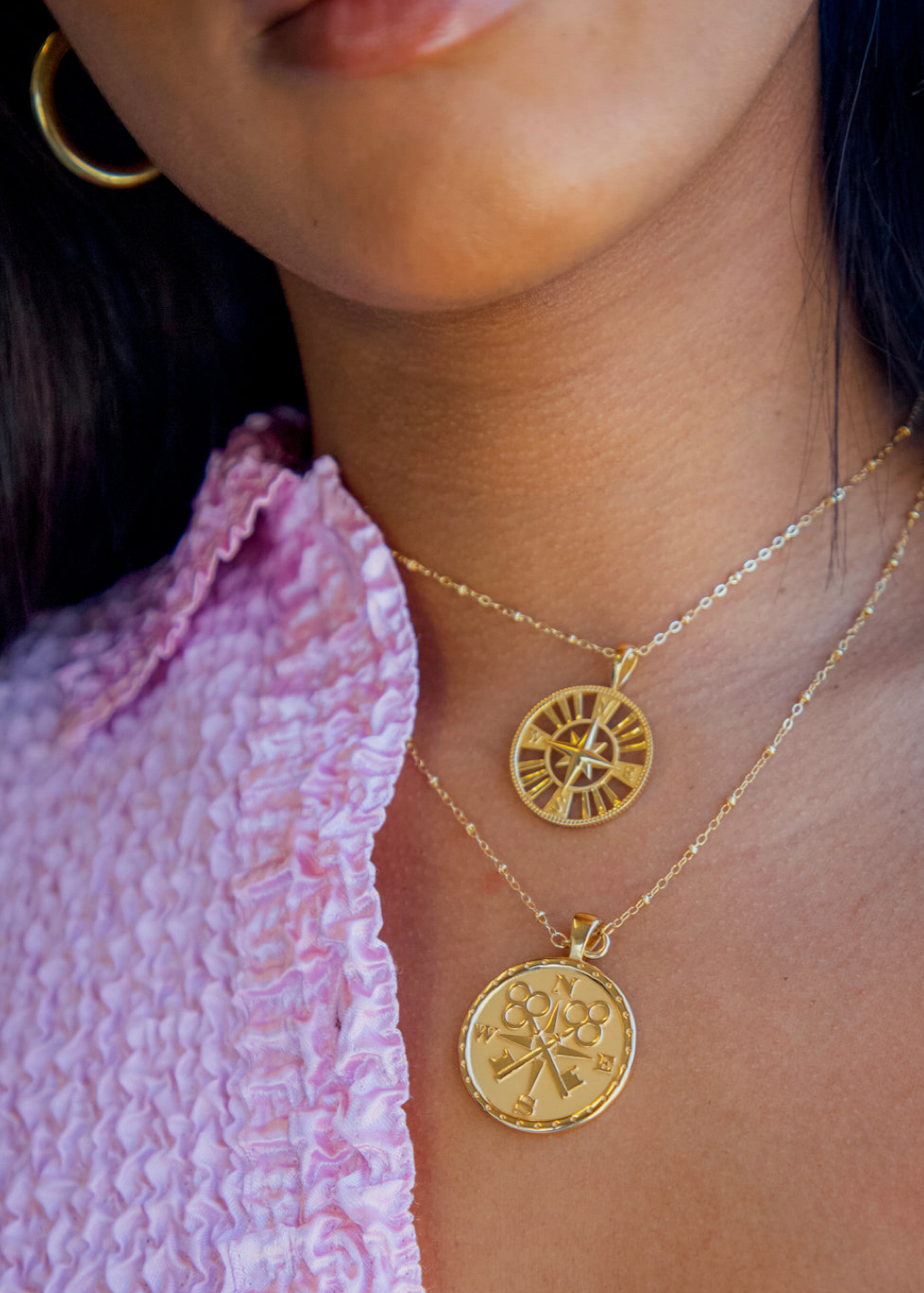 Close-up of a person wearing layered gold necklaces with circular pendant charms, paired with a pink textured top.