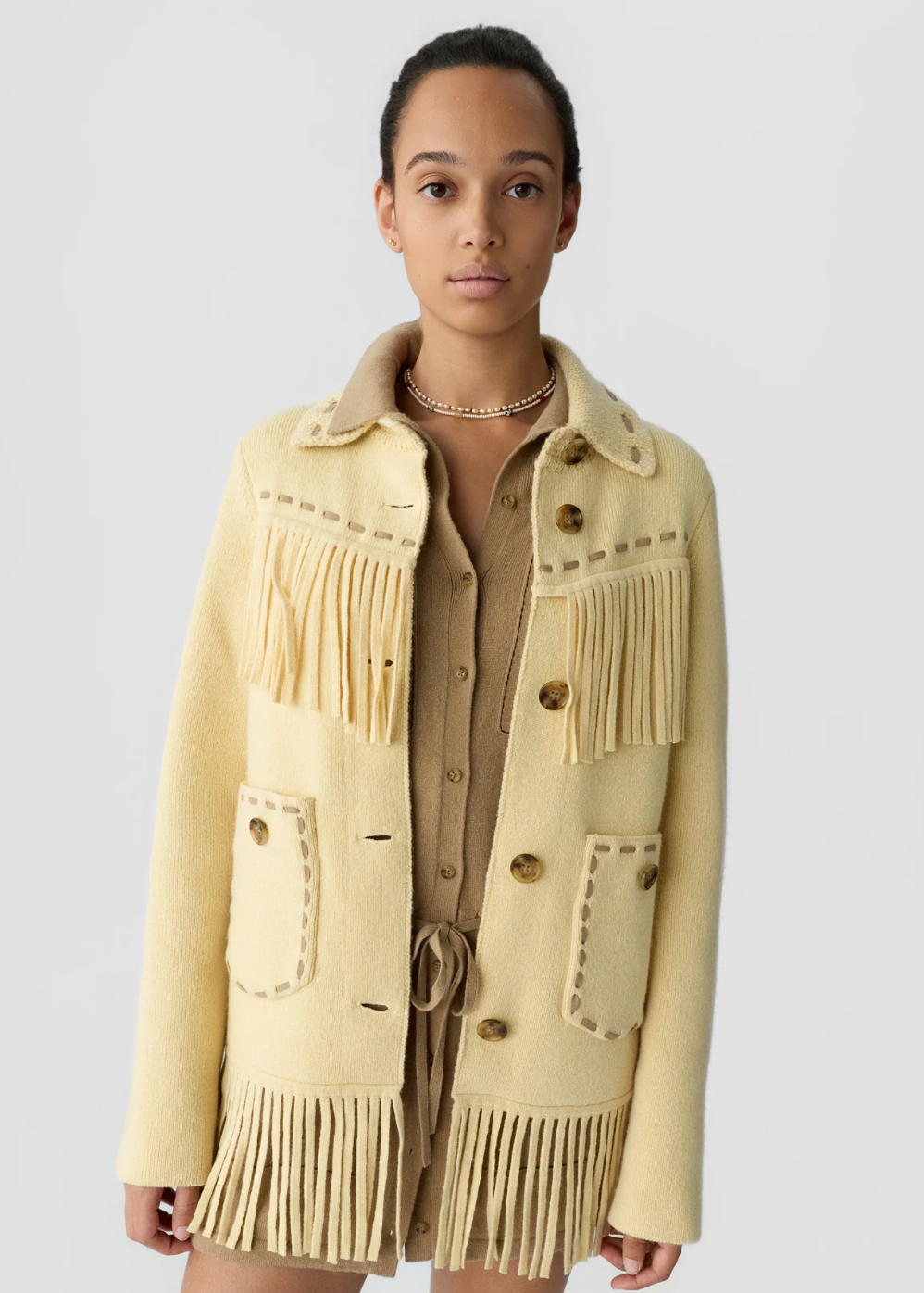 A model wearing a pale yellow fringed knit jacket over a beige button-up shirt, photographed against a solid light gray background.