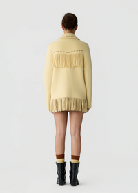 A model viewed from the back wearing a pale yellow knit cardigan with fringe detailing across the shoulders and hem, paired with brown socks and black ankle boots on a light gray studio background.