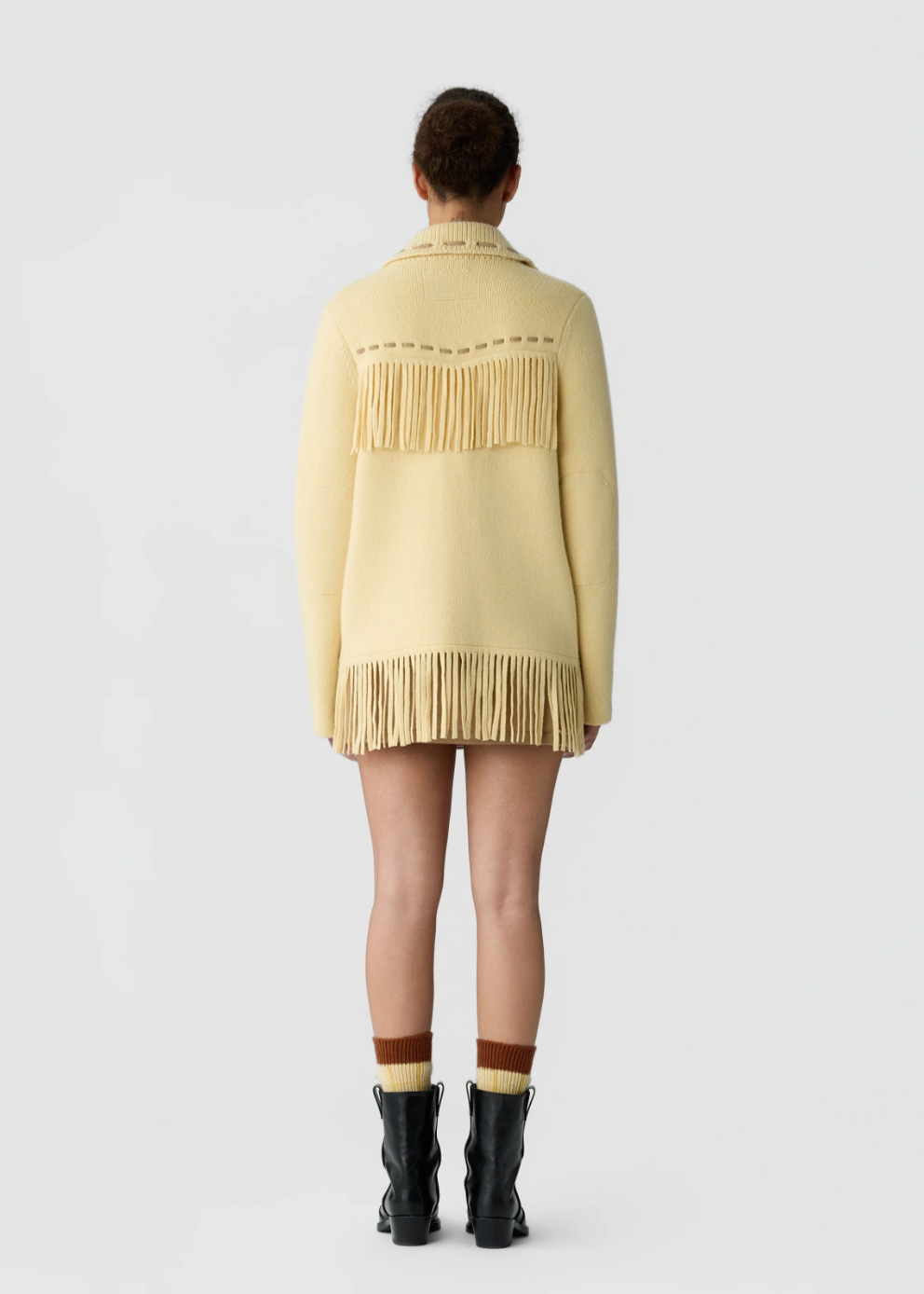 A model viewed from the back wearing a pale yellow knit cardigan with fringe detailing across the shoulders and hem, paired with brown socks and black ankle boots on a light gray studio background.