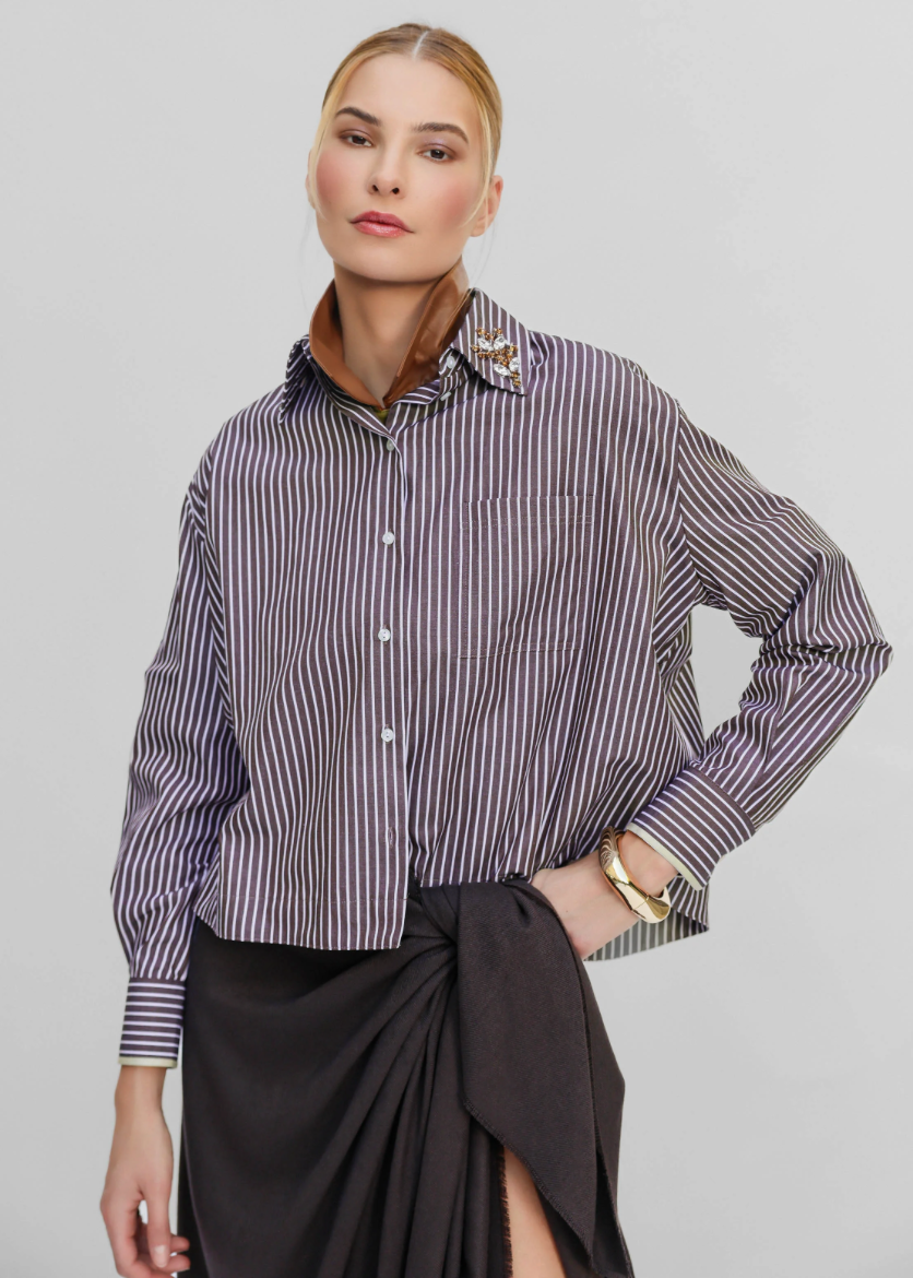 A model wears a long-sleeve button-up shirt with vertical purple and white stripes, featuring a brown collar and decorative gold embroidery on the collar, paired with a dark gray wrap skirt against a light gray background.