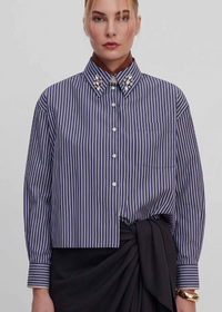 A model wears a navy and white vertically striped button-down shirt with decorative pearl embellishments on the collar, paired with a dark wrap skirt against a light gray studio background.