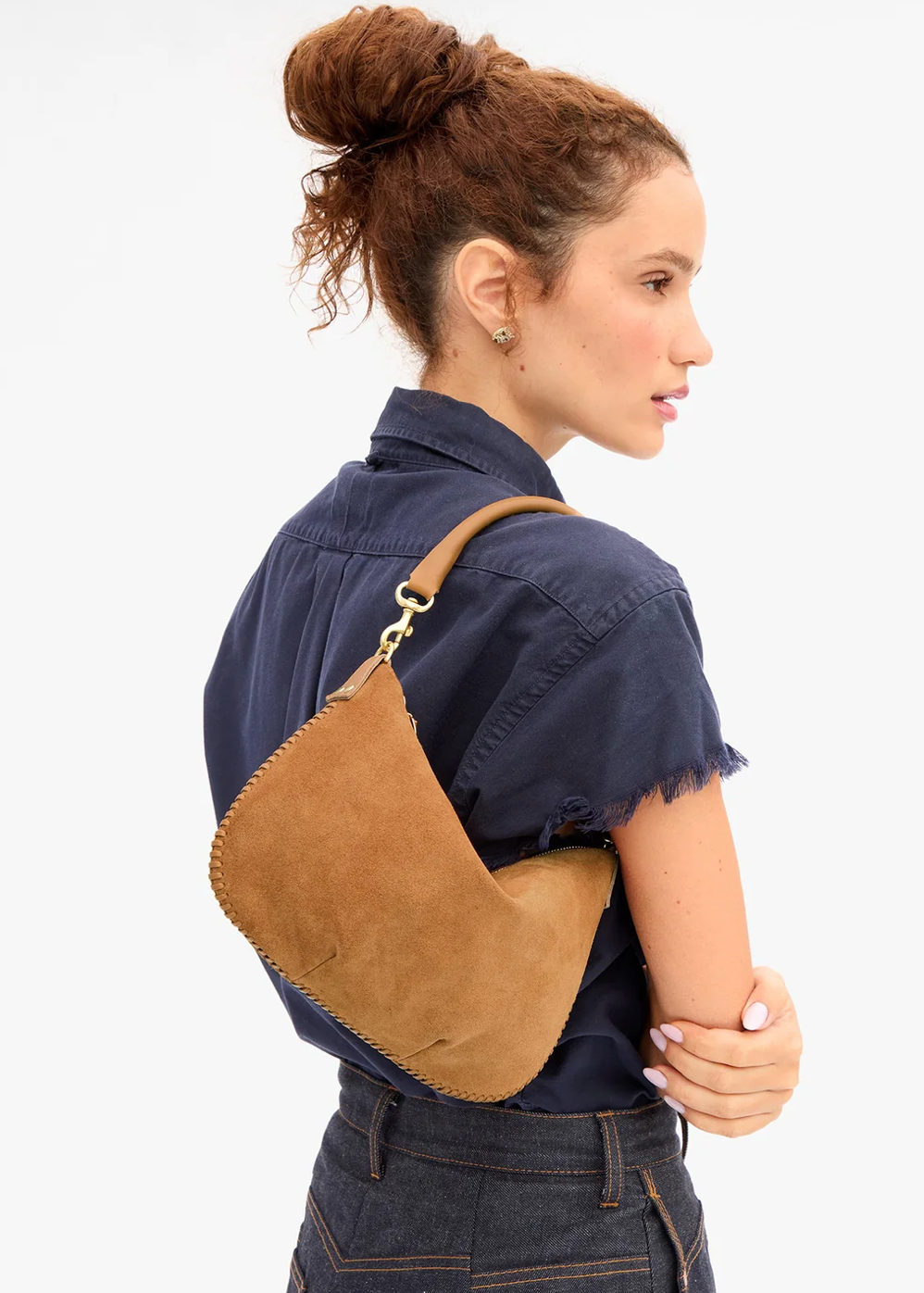 A woman in a navy blue shirt and jeans is shown from the back and side profile, wearing a tan suede crescent-shaped shoulder bag with gold-tone hardware against a plain white background.