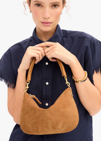 A woman wearing a navy blue shirt holds a tan suede crescent shoulder bag with tan leather handles and gold-tone hardware against a plain white background.