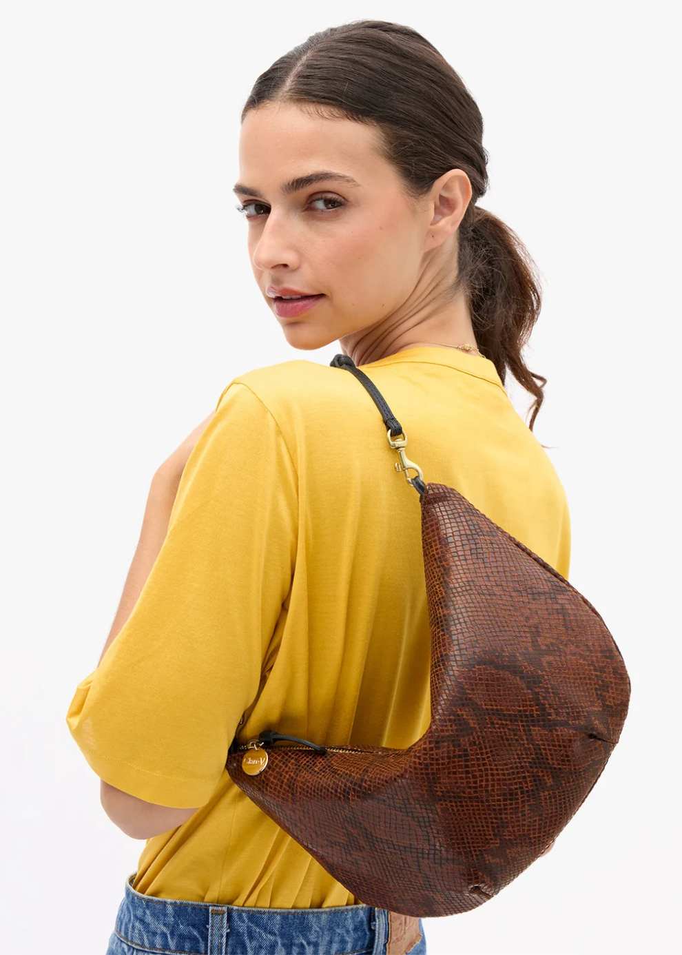 A woman wearing a yellow t-shirt and blue jeans is shown from the back and side, carrying a brown snakeskin-patterned crescent shoulder bag on her shoulder against a plain white background.