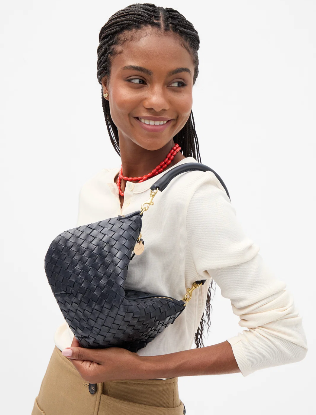 A woman with braided hair models a black woven crescent-shaped shoulder bag with gold-tone hardware and a black strap, wearing a cream sweatshirt and tan pants against a plain white background.