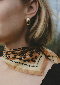 A close-up lifestyle shot of a woman wearing a beige and black leopard-print silk scarf tied around her neck, paired with a gold hoop earring.