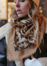A close-up lifestyle shot of a woman wearing a beige scarf with a black and white ornate pattern, styled with a black leather jacket featuring a cream shearling lining.