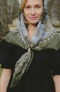 A woman wearing a patterned headscarf tied around her neck with a gold clasp, paired with a black dress, set against a blurred outdoor background.