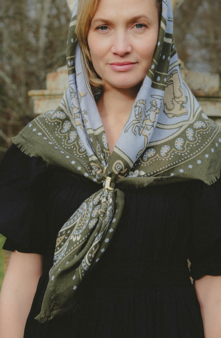 A woman wearing a patterned headscarf tied around her neck with a gold clasp, paired with a black dress, set against a blurred outdoor background.