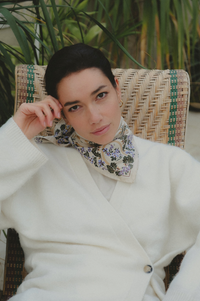 A woman wearing a cream knit cardigan and a floral neck scarf sits in a woven rattan chair with green plants in the background.