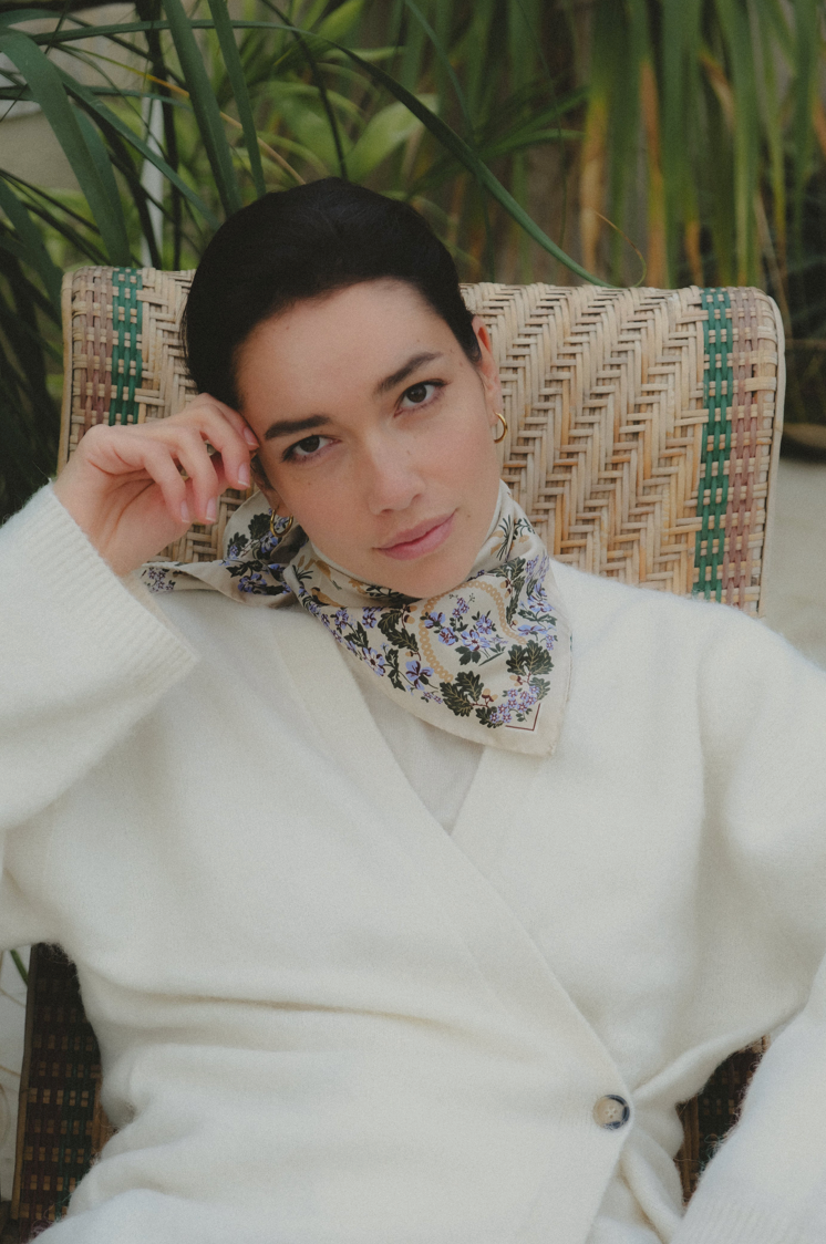 A woman wearing a cream knit cardigan and a floral neck scarf sits in a woven rattan chair with green plants in the background.