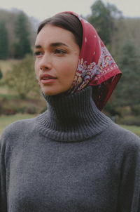 A woman wearing a dark gray ribbed turtleneck sweater and a red paisley patterned headscarf, photographed outdoors against a blurred green forest background.