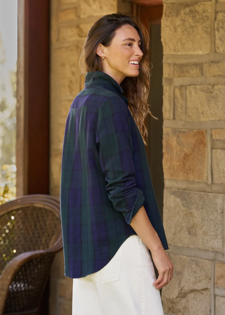 A woman wearing a dark green and navy plaid flannel shirt with white pants, standing in profile in front of a stone wall.