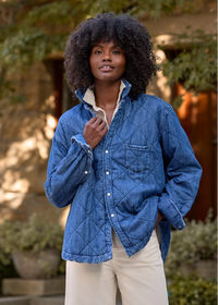 A woman wearing a blue diamond-quilted button-up jacket over a cream shirt and light beige pants, standing outdoors in front of a stone wall and greenery.