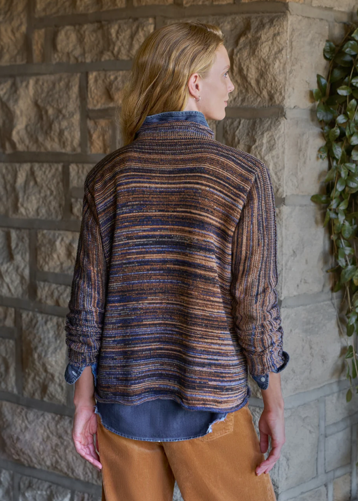 A woman shown from the back wearing a textured, striped knit cardigan in brown, blue, and black, layered over a blue shirt and paired with brown pants, standing against a stone wall with green ivy.