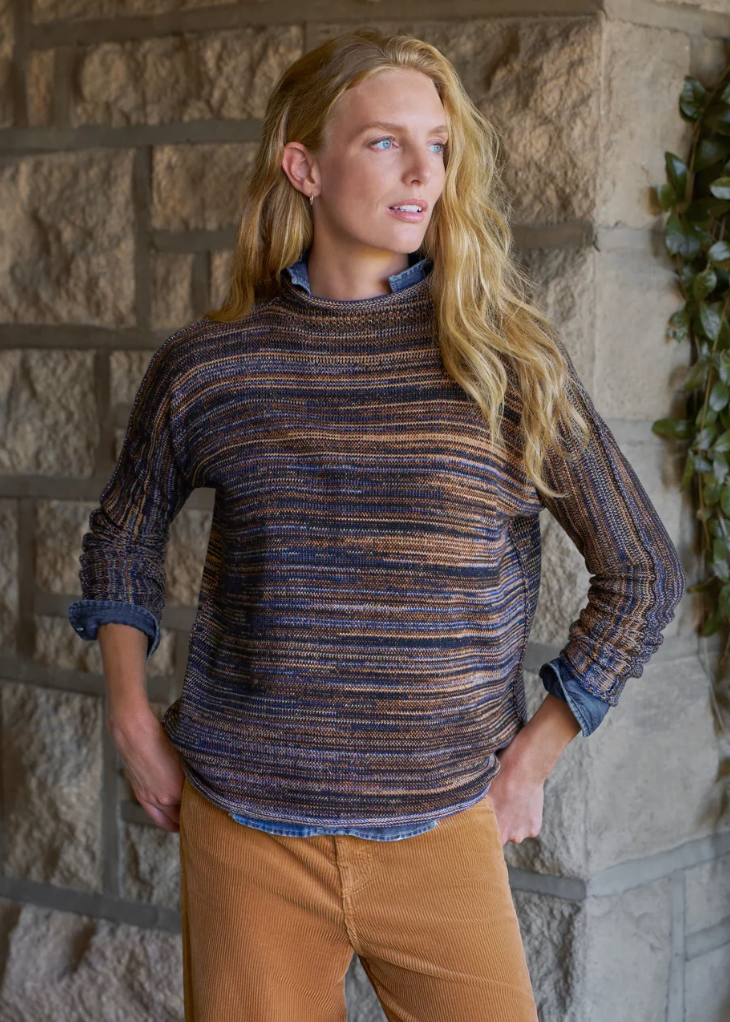 A woman wearing a textured, striped knit mock-neck sweater in navy, blue, and brown tones, layered over a blue denim shirt and paired with tan corduroy pants, standing against a rustic stone wall.