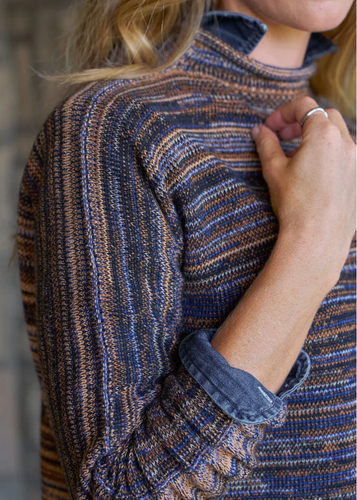Close-up of a person wearing a textured knit mock-neck sweater with horizontal stripes in navy blue, brown, and tan, with a rolled-up denim shirt underneath.