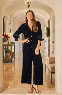 A woman stands in a bright, arched hallway wearing a matching dark navy velvet button-front top and wide-leg pants set, paired with heeled sandals.