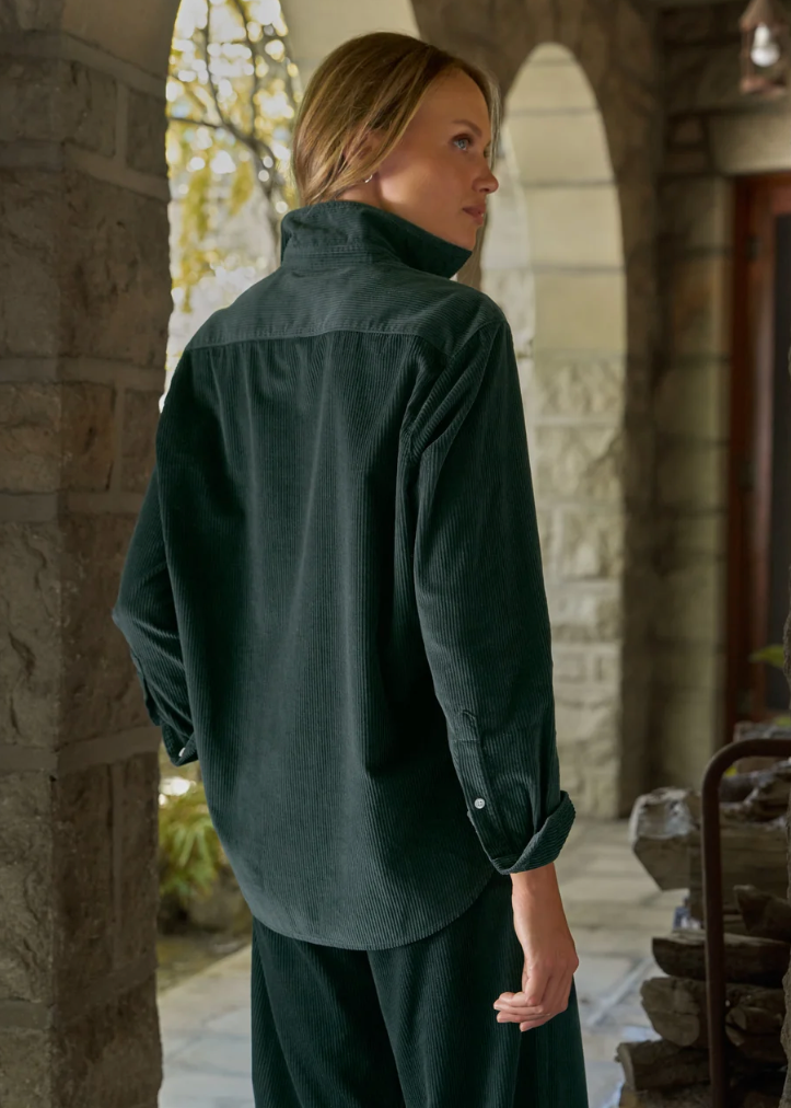 A woman wearing a dark forest green corduroy shirt and matching pants, photographed from behind in a stone archway corridor.