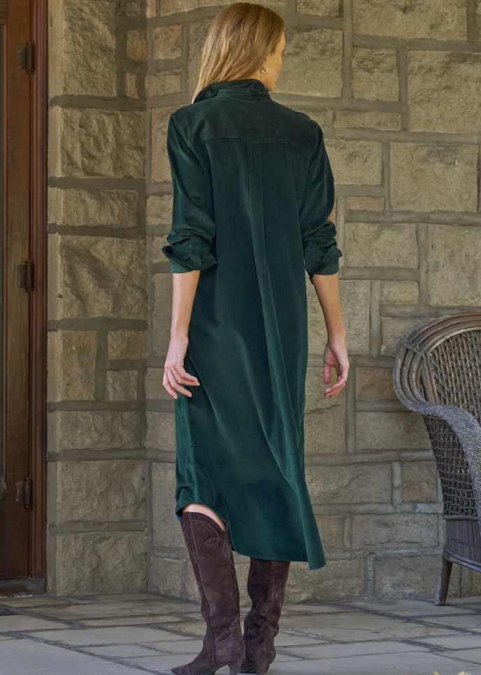 A woman viewed from behind wearing a dark green velvet shirt dress with rolled sleeves and brown over-the-knee boots, standing in front of a stone wall.