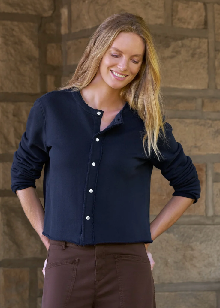 A woman wearing a dark navy long-sleeve button-up shirt and brown pants, standing in front of a rustic stone wall.