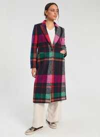 A model stands against a light gray studio background wearing a long-line plaid coat with a vibrant mix of pink, green, black, and red stripes, paired with a white t-shirt, cream wide-leg trousers, and brown sneakers.