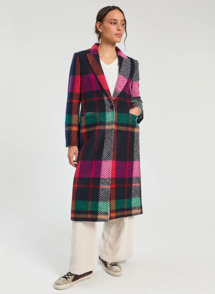 A model stands against a light gray studio background wearing a long-line plaid coat with a vibrant mix of pink, green, black, and red stripes, paired with a white t-shirt, cream wide-leg trousers, and brown sneakers.